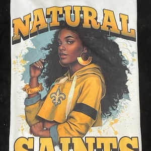 Puede incluir: Camiseta blanca con un estampado gráfico de una mujer de piel morena y cabello rizado que lleva una sudadera con capucha amarilla y negra con el logotipo de los New Orleans Saints. El texto "NATURAL SAINTS Girl" está impreso en amarillo y blanco.