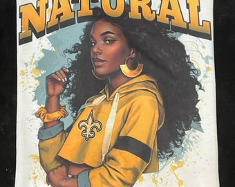 May include: White t-shirt with a graphic print of a woman with brown skin and curly hair wearing a yellow and black hoodie with the New Orleans Saints logo. The text "NATURAL SAINTS Girl" is printed in yellow and white.