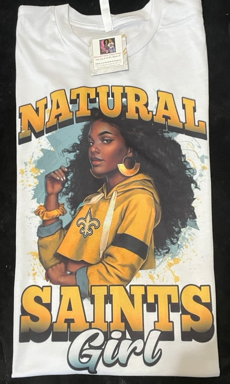 May include: White t-shirt with a graphic print of a woman with brown skin and curly hair wearing a yellow and black hoodie with the New Orleans Saints logo. The text "NATURAL SAINTS Girl" is printed in yellow and white.