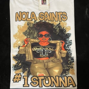 Puede incluir: Camiseta blanca con un gráfico dorado y negro de una mujer con gafas de sol y una chaqueta con el texto "Who Dat Chic" y el logotipo de los New Orleans Saints. El texto "NOLA SAINTS" está en la parte superior de la camiseta y "#1 STUNNA" en la parte inferior.