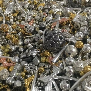 May include: A large assortment of silver, gold, and black metal beads and charms. The beads are of various shapes and sizes, including round, square, and heart-shaped. Some of the charms are in the shape of animals, flowers, and other designs.