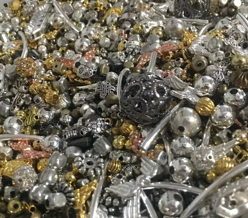 May include: A large assortment of silver, gold, and black metal beads and charms. The beads are of various shapes and sizes, including round, square, and heart-shaped. Some of the charms are in the shape of animals, flowers, and other designs.