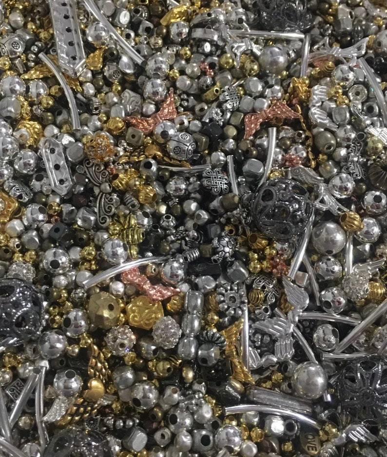 May include: A large assortment of silver, gold, and black beads and charms. The beads are of various shapes and sizes, including round, square, and heart-shaped. The charms are also of various shapes and sizes, including wings, flowers, and stars.