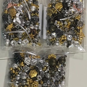 May include: Three bags of assorted silver, gold, and bronze beads. The beads are in various shapes and sizes, including round, square, and star-shaped.