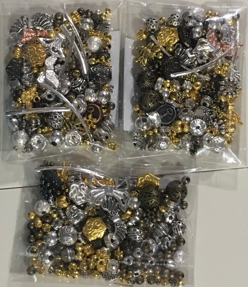 May include: Three bags of assorted silver, gold, and bronze beads. The beads are in various shapes and sizes, including round, square, and star-shaped.