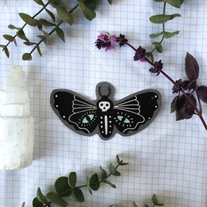 May include: A black moth-shaped patch with a skull and skeleton design, featuring glowing green eyes. The patch is set against a white grid background, with sprigs of greenery and a white crystal formation. The moth's wings have white accents.
