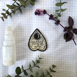 May include: A cream-colored, heart-shaped patch with the text "AS ABOVE SO BELOW" and a crescent moon design. The patch is surrounded by sprigs of greenery and a white selenite crystal tower on a grid background.