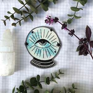 May include: A patch featuring a stylized fortune-telling crystal ball with an eye design. The crystal ball has a light blue and white watercolor effect, surrounded by radiating lines and black stars. The patch is on a white grid background with green and purple foliage.