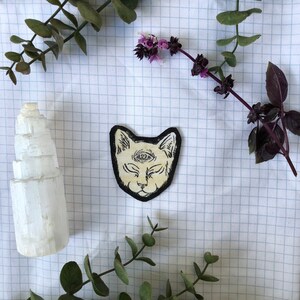 May include: A cream-colored cat patch with a third eye design, outlined in black, rests on a grid-patterned surface. The patch is surrounded by sprigs of greenery and a white, layered crystal formation. The image has a natural, bohemian aesthetic.