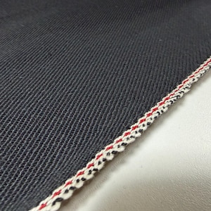 Puede incluir: Primer plano de tela vaquera azul oscuro con un tejido texturizado. El borde est&aacute; acabado con un ribete decorativo con hilos rojos, blancos y negros, creando un patr&oacute;n de rayas. La tela es probablemente para ropa.