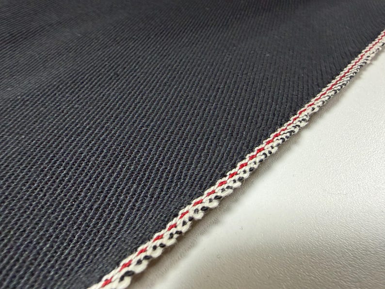 Puede incluir: Primer plano de tela vaquera azul oscuro con un tejido texturizado. El borde est&aacute; acabado con un ribete decorativo con hilos rojos, blancos y negros, creando un patr&oacute;n de rayas. La tela es probablemente para ropa.