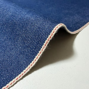 Könnte beinhalten: Ein Stück tiefblauer Denim-Stoff mit einer ausgeprägten Webkante. Der Stoff weist eine sichtbare Köperbindung auf und hat einen sauberen, verarbeiteten Rand, der durch einen dünnen, leuchtend roten Faden entlang der weißen Webkante gekennzeichnet ist.
