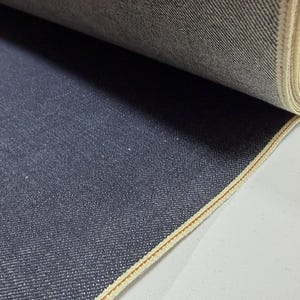 May include: A roll of dark blue denim fabric. The fabric has a textured surface with a visible weave. The edge of the fabric is finished with a light beige selvedge. The fabric is tightly rolled.