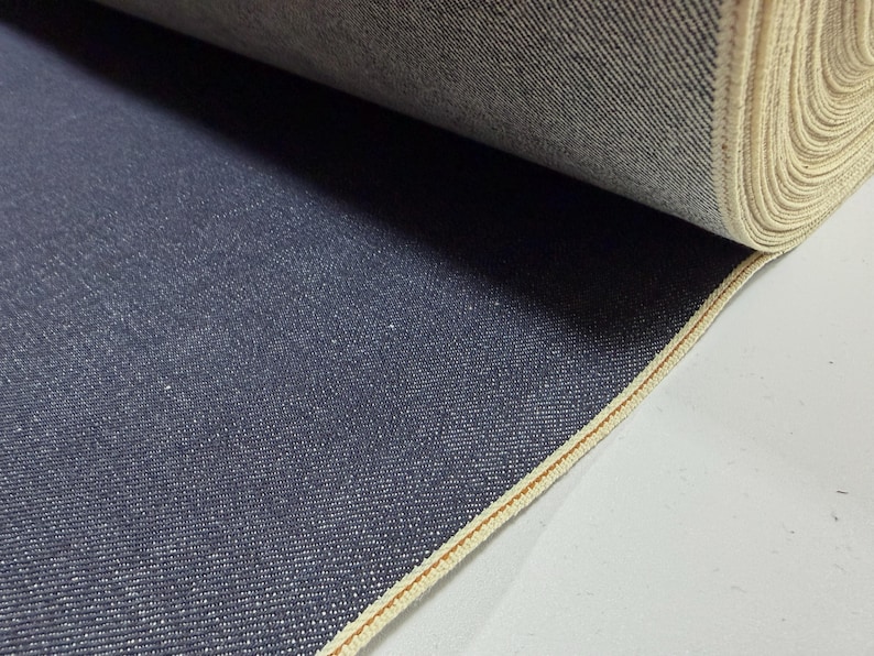 May include: A roll of dark blue denim fabric. The fabric has a textured surface with a visible weave. The edge of the fabric is finished with a light beige selvedge. The fabric is tightly rolled.