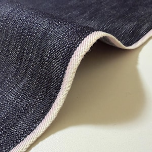 Könnte beinhalten: Nahaufnahme eines dunkelblauen Denim-Stoffs mit weißem und hellrosa Besatz. Der Stoff ist strukturiert und gefaltet, wodurch das Gewebe des Materials sichtbar wird. Der Rand ist gebogen und zeigt die Dicke des Stoffes.