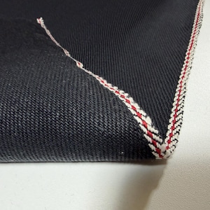 Puede incluir: Primer plano de una esquina de tela vaquera oscura. La tela tiene un tejido texturizado y est&aacute; rematada con un borde cosido rojo, blanco y negro. La esquina est&aacute; doblada, revelando las costuras contrastantes.