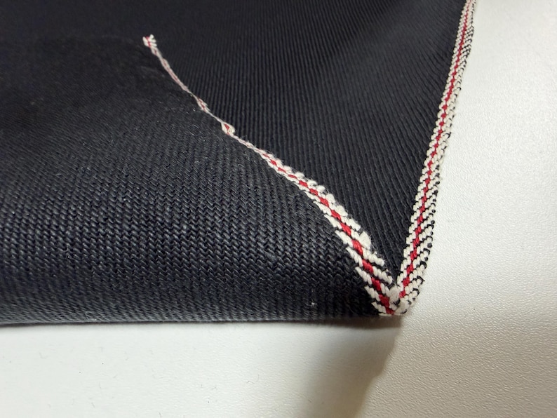 Puede incluir: Primer plano de una esquina de tela vaquera oscura. La tela tiene un tejido texturizado y est&aacute; rematada con un borde cosido rojo, blanco y negro. La esquina est&aacute; doblada, revelando las costuras contrastantes.