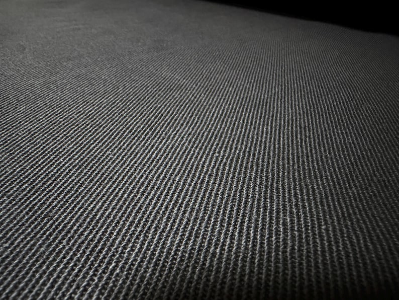 Puede incluir: Primer plano de una tela gris oscuro texturizada con un patr&oacute;n acanalado. La tela parece estar tejida, con l&iacute;neas verticales visibles y una superficie ligeramente irregular, creando una sensaci&oacute;n de profundidad y textura. La iluminaci&oacute;n resalta el tejido.