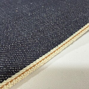 May include: Close-up of dark blue denim fabric with white flecks. The edge of the fabric is finished with a cream-colored selvedge and a thin brown stripe. The fabric has a textured appearance.