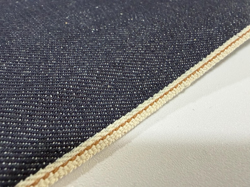 May include: Close-up of dark blue denim fabric with white flecks. The edge of the fabric is finished with a cream-colored selvedge and a thin brown stripe. The fabric has a textured appearance.