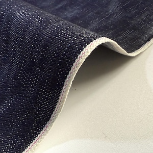 Może przedstawiać: Zbliżenie ciemnoniebieskiego materiału dżinsowego z białym brzegiem. Tkanina ma teksturowany wygląd z małymi białymi plamkami. Krawędź wykończona jest białą i różową nicią.