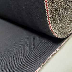 Puede incluir: Un rollo de tela vaquera gris oscuro. La tela tiene un patr&oacute;n de tejido diagonal y un borde de orillo con rayas rojas y blancas. El rollo est&aacute; bien enrollado, mostrando las variaciones de textura y color del material.