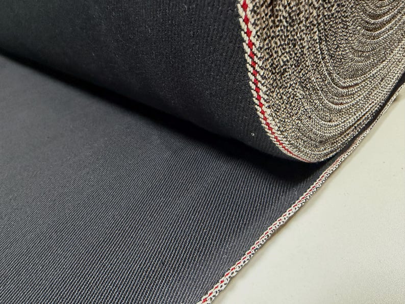 Puede incluir: Un rollo de tela vaquera gris oscuro. La tela tiene un patr&oacute;n de tejido diagonal y un borde de orillo con rayas rojas y blancas. El rollo est&aacute; bien enrollado, mostrando las variaciones de textura y color del material.