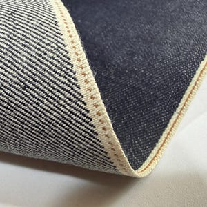 May include: Close-up of denim fabric with a white and black diagonal weave. The edge of the fabric is visible, showing a beige selvedge with a brown stitch. The dark blue denim is folded over, revealing the interior texture.