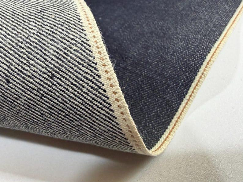 May include: Close-up of denim fabric with a white and black diagonal weave. The edge of the fabric is visible, showing a beige selvedge with a brown stitch. The dark blue denim is folded over, revealing the interior texture.