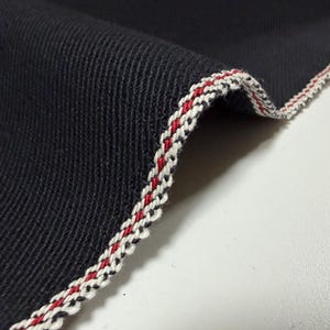 Puede incluir: Primer plano de una tela azul marino oscuro con un borde decorativo. El borde presenta un ribete trenzado en blanco, rojo y negro, que a&ntilde;ade un detalle de contraste al textil. La textura de la tela es visible.