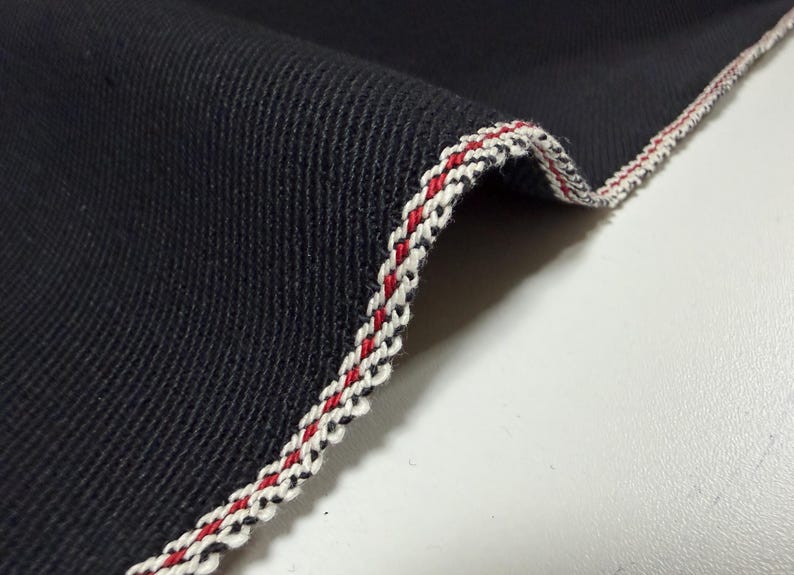Puede incluir: Primer plano de una tela azul marino oscuro con un borde decorativo. El borde presenta un ribete trenzado en blanco, rojo y negro, que a&ntilde;ade un detalle de contraste al textil. La textura de la tela es visible.