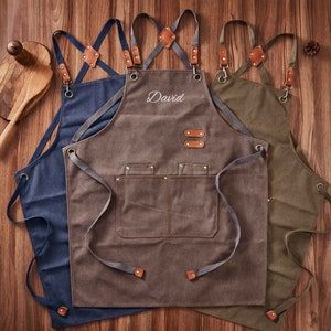 May include: Three canvas aprons with adjustable straps. The centre apron is brown with the name "David" embroidered on the front. The apron on the left is blue and the apron on the right is green.