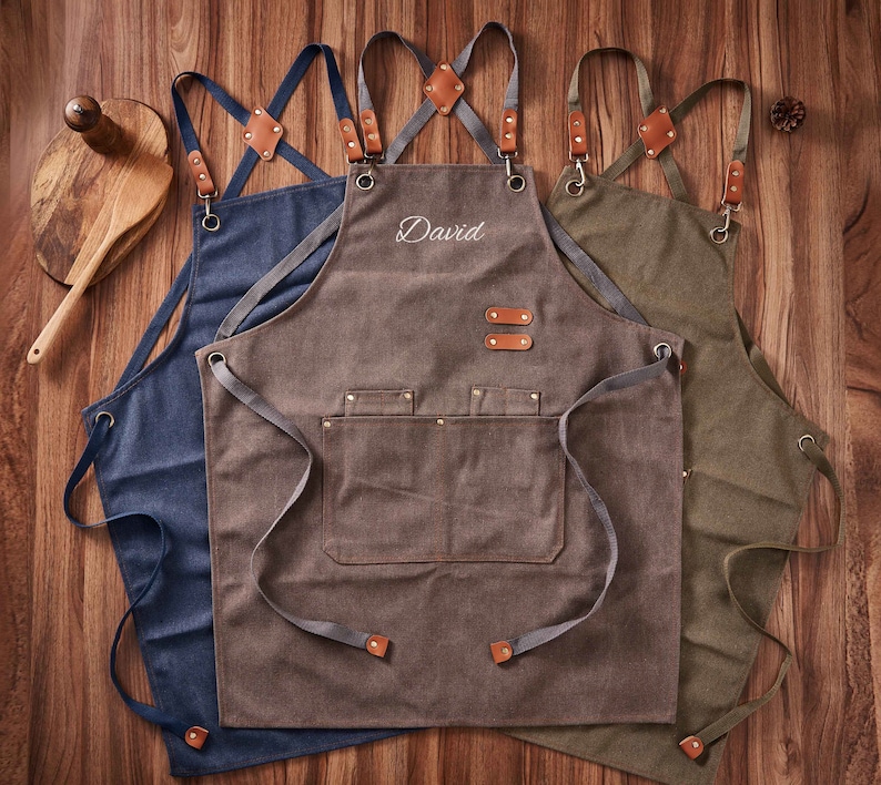 May include: Three canvas aprons with adjustable straps. The centre apron is brown with the name "David" embroidered on the front. The apron on the left is blue and the apron on the right is green.