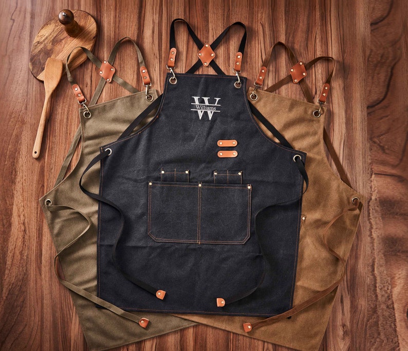 May include: Three denim aprons with adjustable straps and multiple pockets. The centre apron has the name "Williams" embroidered on it.