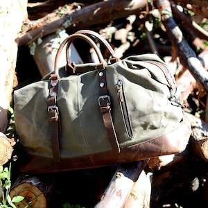 May include: An olive green canvas duffel bag with brown leather accents. The bag features a brown leather base, handles, and straps with silver buckles. A black zipper runs down the side. The bag is designed for travel.