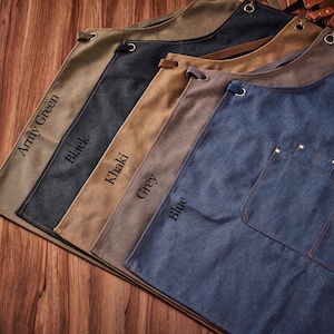 May include: Five canvas aprons in different colours, featuring leather straps and metal rivets. The colours are army green, black, khaki, grey, and blue. The aprons are arranged in a row from left to right, with the army green apron at the top and the blue apron at the bottom.