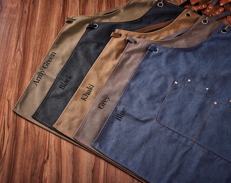 May include: Five canvas aprons in different colours, featuring leather straps and metal rivets. The colours are army green, black, khaki, grey, and blue. The aprons are arranged in a row from left to right, with the army green apron at the top and the blue apron at the bottom.