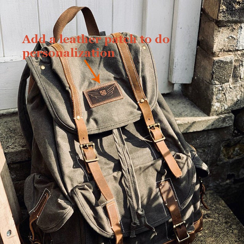 Puede incluir: Una mochila de lona verde con correas y hebillas de cuero marr&oacute;n. La mochila tiene un peque&ntilde;o parche de cuero con un logotipo en la parte delantera. El texto "A&ntilde;adir un parche de cuero para hacer la personalizaci&oacute;n" est&aacute; escrito en la imagen.