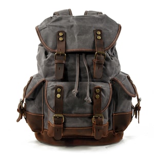 Puede incluir: Una mochila de lona gris con correas y hebillas de cuero marr&oacute;n. La mochila tiene un cierre de cord&oacute;n y dos bolsillos grandes en la parte delantera.