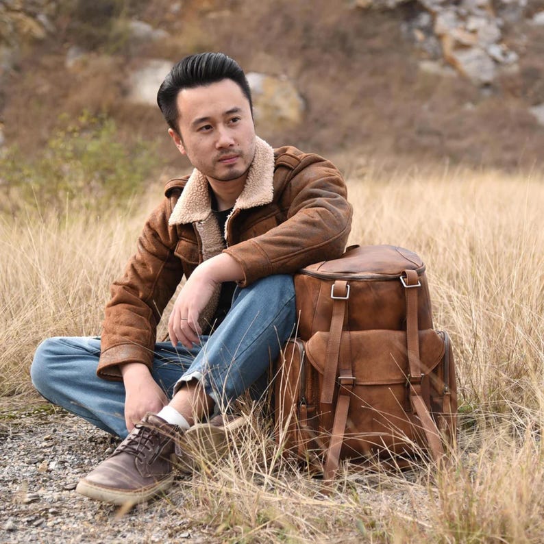 May include: A brown leather backpack with multiple compartments and adjustable straps. The backpack has a vintage look with metal buckles and a flap closure. The backpack is sitting on the ground next to a person wearing a brown jacket.