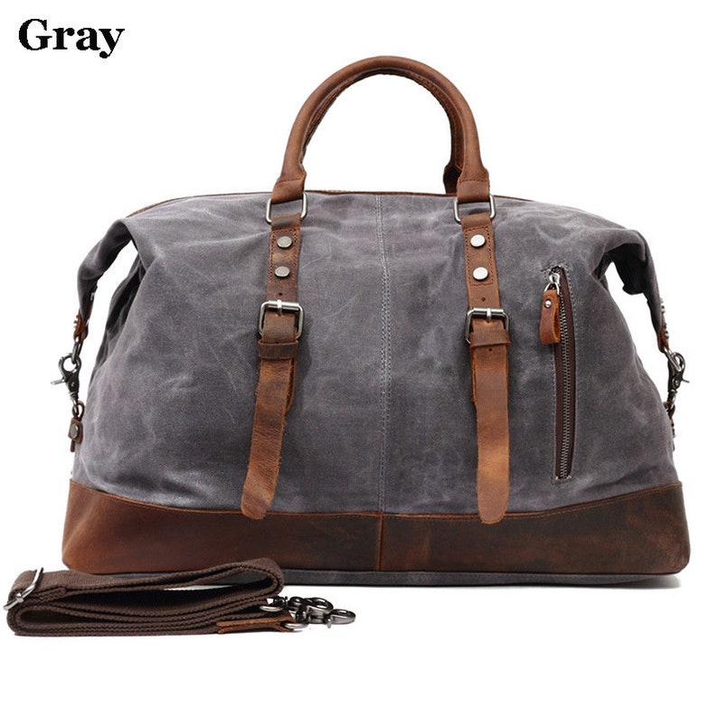 May include: Gray canvas duffel bag with brown leather accents. Features include a brown leather base, handles, and straps with silver buckles. Includes a detachable shoulder strap and a side zipper pocket. The word "Gray" is in the top left corner.