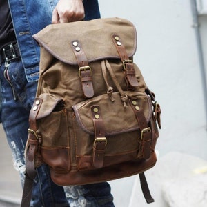 Puede incluir: Una mochila de lona marr&oacute;n con detalles de cuero y m&uacute;ltiples hebillas. La mochila tiene un compartimento principal grande y dos bolsillos delanteros m&aacute;s peque&ntilde;os.