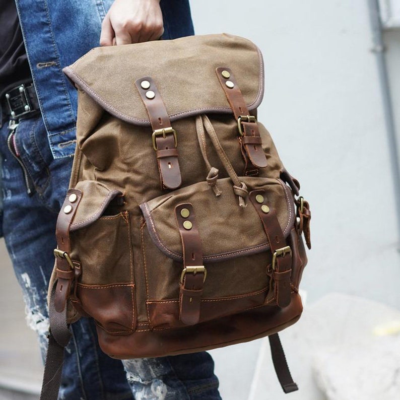 Puede incluir: Una mochila de lona marr&oacute;n con detalles de cuero y m&uacute;ltiples hebillas. La mochila tiene un compartimento principal grande y dos bolsillos delanteros m&aacute;s peque&ntilde;os.
