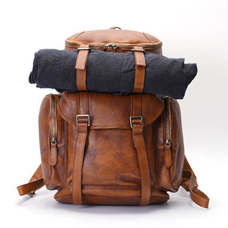 May include: A brown leather backpack with a rolled-up black blanket secured on top. The backpack has multiple compartments, adjustable straps, and silver buckles. The leather has a distressed finish, giving it a vintage look.