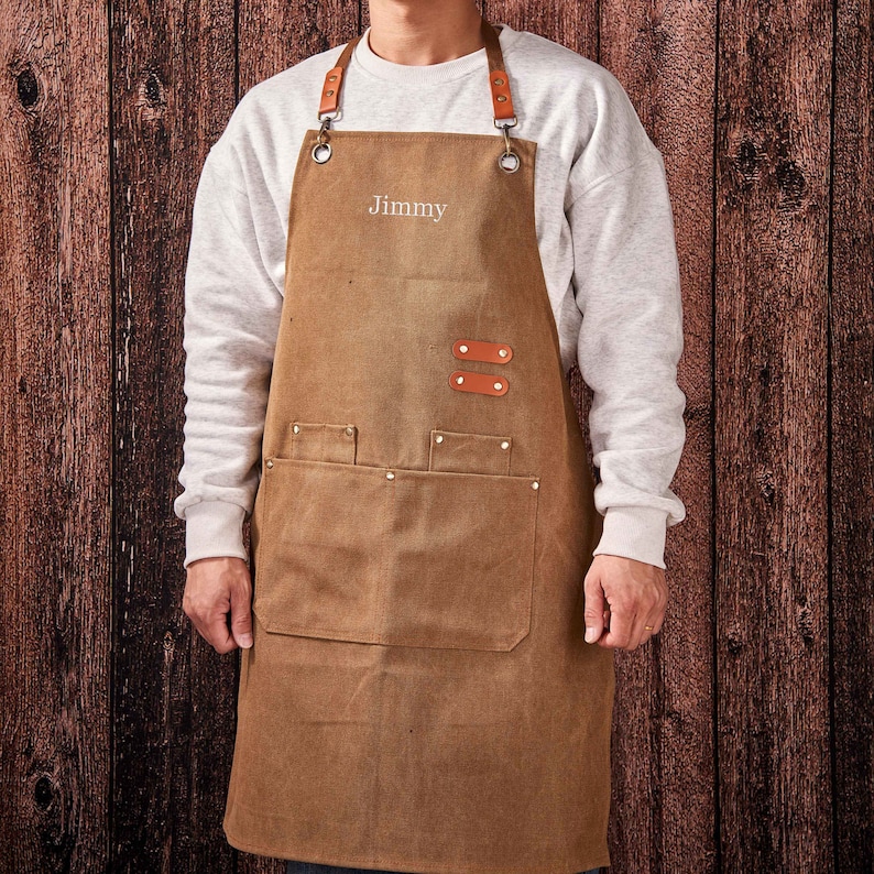 May include: A brown canvas apron with leather straps and a large pocket. The apron has the name "Jimmy" embroidered on the front.