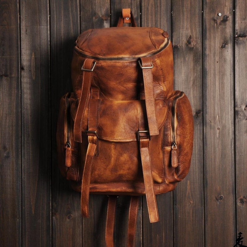 May include: A brown leather backpack with a vintage, distressed appearance. The backpack features multiple compartments, including side pockets with zippers and a front flap secured by buckles. Adjustable straps and a top handle are also visible.