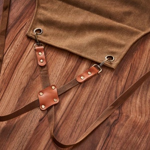 May include: Brown canvas apron with adjustable straps and leather accents. The straps have metal buckles and are secured with rivets. The apron is on a wooden surface.