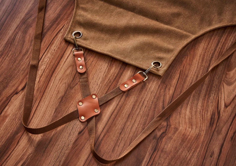 May include: Brown canvas apron with adjustable straps and leather accents. The straps have metal buckles and are secured with rivets. The apron is on a wooden surface.