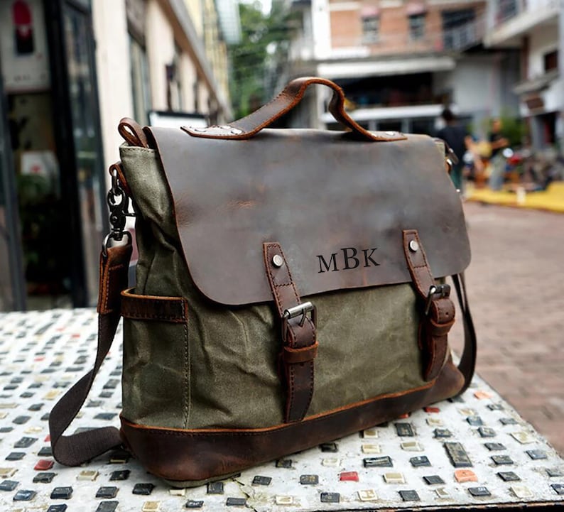 May include: A green canvas messenger bag with a brown leather flap and accents. The bag features a shoulder strap, a top handle, and the initials "MBK" embossed on the front. The bag has a vintage, rugged aesthetic.