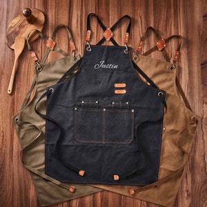 May include: Three brown canvas aprons with adjustable straps. The centre apron is black denim with a large pocket and the name "Justin" embroidered on the front.